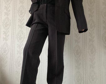 Op de afbeelding: Een donkerbruin pak bestaande uit een blazer en een broek met wijde pijpen. De blazer heeft een revers met inkeping en een knoopsluiting. De broek is hoog getailleerd. Een zwarte trui en een goudkleurige ketting zijn zichtbaar. De outfit wordt gepresenteerd tegen een achtergrond met patroon.