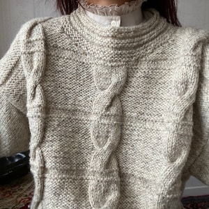 May include: A light beige, hand-knitted jumper with a high, ribbed collar and cable knit detailing. The jumper features a textured pattern with horizontal stripes and vertical cable designs. The sleeves are long and have a slightly puffed shoulder design.