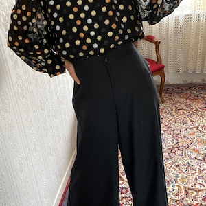 May include: A black sheer blouse with gold, white, and brown polka dots. The blouse has long sleeves and a round neckline. The model is wearing the blouse with black wide-leg pants.