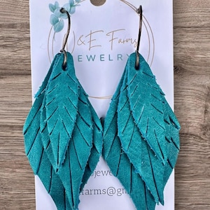 May include: Teal leather feather earrings. Each earring features three layered, leaf-shaped pieces with detailed cut lines. The earrings hang from bronze-colored ear wires. The earrings are displayed on a white card with the text "J & E Farms JEWELRY".