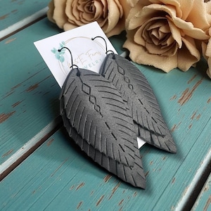 May include: A pair of dark gray leaf-shaped earrings with intricate etched details. The earrings are made of a textured material and hang from black ear wires. The earrings are displayed on a turquoise surface with a floral background.