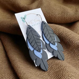 May include: Gray and light blue leaf-shaped earrings. The earrings are made of layered leather, with a textured gray top layer and a light blue layer underneath. The earrings hang from silver-colored hoops.