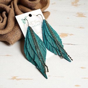 May include: Teal leather feather earrings with bronze-colored chain accents. The earrings have a layered design and are attached to bronze-colored ear wires. The earrings are displayed on a white card with the text "W & E Fairy WELRY".
