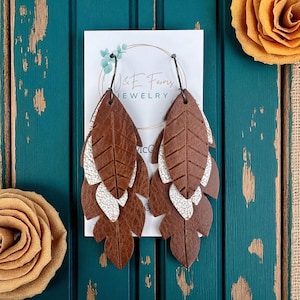 May include: Brown leather leaf earrings with white textured accents. The earrings are layered and feature a leaf design. The earrings are hanging on a white card with the text "J&E Farms JEWELRY".