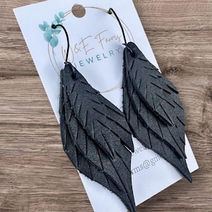 May include: Black leather feather earrings with detailed veining. The earrings are hanging from black ear wires. The earrings are displayed on a white card with the text "W & E Farms Jewelry".