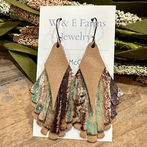 May include: A pair of brown leather earrings with a teal and brown patterned design. The earrings are shaped like a teardrop and have a fringe-like design. The earrings are labeled "W & E Farms Jewelry".