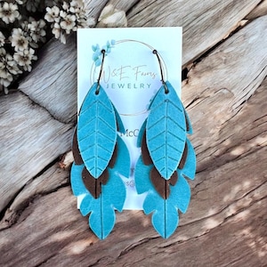 May include: Turquoise and brown leather leaf earrings. The earrings are layered with three leaf shapes in varying sizes. The top and bottom leaves are turquoise, and the middle leaf is brown. The earrings hang from a black wire hook. The earrings are displayed on a white card with the text "W&E Farms JEWELRY".