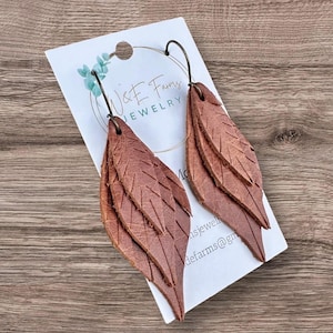 May include: A pair of brown leather leaf earrings with bronze-colored ear wires. The earrings are layered with detailed veining and are displayed on a white card with the text "W&E Farms JEWELRY".