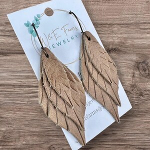 May include: A pair of tan leather earrings featuring a layered, feather-like design with etched lines. The earrings are attached to a white display card printed with the text W&E Farms JEWELRY, featuring a small blue eucalyptus leaf graphic.