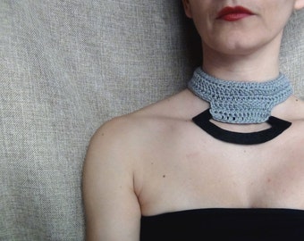 May include: A woman wearing a gray crocheted choker necklace with a black felt accent. The necklace has a geometric shape and is worn against a gray fabric background.