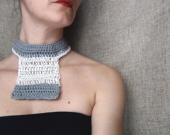 May include: A woman wearing a gray and white crocheted collar necklace. The necklace is made of two different types of yarn, one gray and one white. The woman is wearing a black top and has her hair pulled back.
