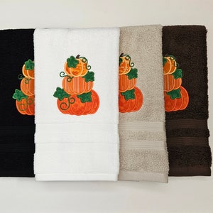 May include: Four bath towels in black, white, tan, and brown, each embroidered with a stack of three orange pumpkins with green leaves. The towels are arranged side by side against a white background, showcasing a fall or autumn theme.