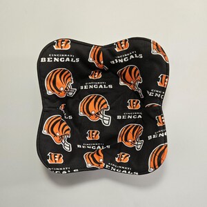 May include: A black, padded item with a repeating pattern of orange and black Cincinnati Bengals football helmets and the team name. The item has a curved shape and is likely a kitchen accessory.