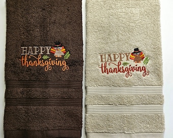 May include: Two rectangular Thanksgiving hand towels. One is dark brown, the other is beige. Both have the embroidered text "HAPPY thanksgiving" with a turkey and fall leaves design. The towels are made of a soft, absorbent material.