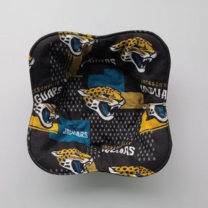 May include: A square, fabric bowl cozy featuring a repeating pattern of Jacksonville Jaguars logos and jaguar heads in teal, gold, and black. The text "Jaguars" is visible. The cozy is designed to hold a bowl of food.