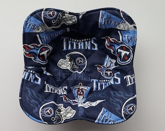 May include: A navy blue bowl cozy featuring a repeating pattern of the Tennessee Titans logo and helmet in light blue, red, and white. The cozy is quilted and has a curved shape.