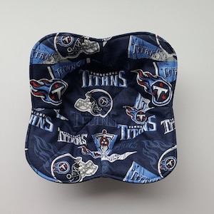 May include: A navy blue bowl cosy with a repeating pattern of the Tennessee Titans logo and helmet in light blue, red, and white. The cosy is quilted and has a curved shape.