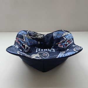 May include: A navy blue bowl cosy with a printed pattern featuring the Tennessee Titans logo and team name in white, light blue, and red. The bowl has a square shape with a folded rim.