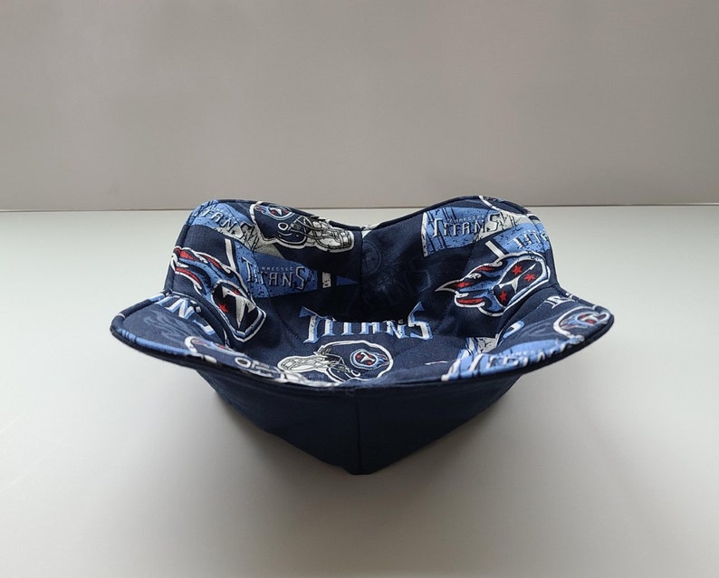 May include: A navy blue bowl cosy with a printed pattern featuring the Tennessee Titans logo and team name in white, light blue, and red. The bowl has a square shape with a folded rim.