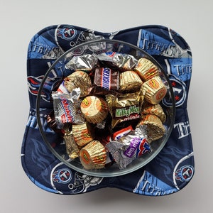 May include: A clear glass bowl filled with assorted wrapped sweets, resting on a navy blue and light blue padded bowl holder. The bowl holder features a repeating pattern of the Tennessee Titans logo and team name.