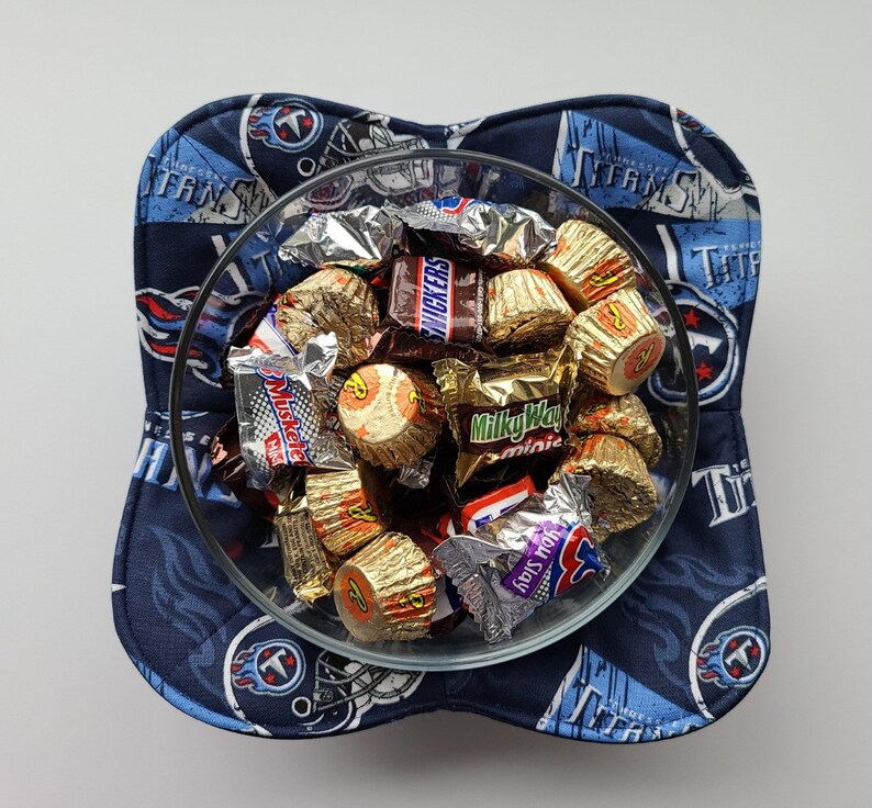 May include: A clear glass bowl filled with assorted wrapped sweets, resting on a navy blue and light blue padded bowl holder. The bowl holder features a repeating pattern of the Tennessee Titans logo and team name.