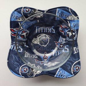 May include: A clear glass bowl sits inside a navy blue bowl cosy. The cosy is patterned with the words "Tennessee Titans", team logos, and helmets in white, light blue, and red. The bowl cosy is square with rounded corners.
