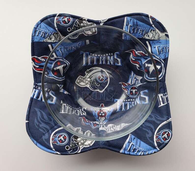 May include: A clear glass bowl sits inside a navy blue bowl cosy. The cosy is patterned with the words "Tennessee Titans", team logos, and helmets in white, light blue, and red. The bowl cosy is square with rounded corners.