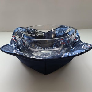 May include: A clear glass bowl sits inside a navy blue fabric bowl cosy. The cosy has a blue and white patterned fabric with the words "TITANS" and team logos. The cosy is square-shaped with folded corners.