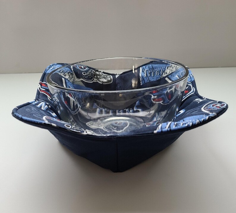 May include: A clear glass bowl sits inside a navy blue fabric bowl cosy. The cosy has a blue and white patterned fabric with the words "TITANS" and team logos. The cosy is square-shaped with folded corners.