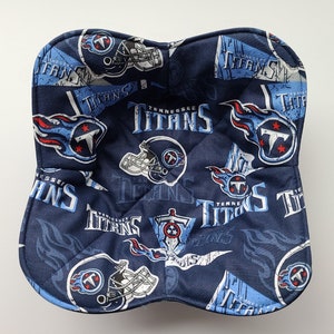 May include: A navy blue bowl cosy with a repeating pattern of the Tennessee Titans logo, helmets, and the team name in white and light blue. The cosy is quilted and has a square shape with rounded corners.