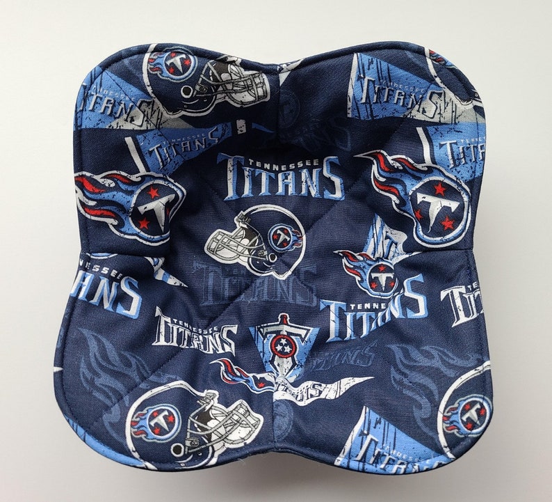 May include: A navy blue bowl cosy with a repeating pattern of the Tennessee Titans logo, helmets, and the team name in white and light blue. The cosy is quilted and has a square shape with rounded corners.