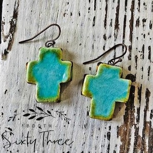 May include: A pair of turquoise cross earrings with a green and brown trim. The earrings have a rustic, handmade appearance with dark metal hooks. The image is set against a weathered, white wooden background.