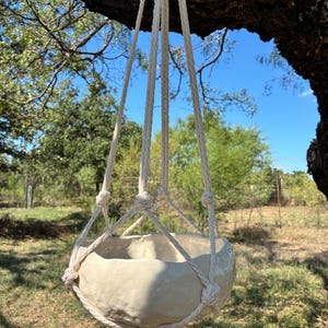 May include: A cream-colored ceramic bowl with an irregular, organic shape suspended by a white macrame rope hanger. The hanger features knotted cords that cradle the bowl, extending upward to a single point of suspension.
