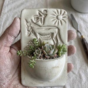 May include: A rectangular off-white ceramic wall planter featuring a relief carving of a donkey, a bird, and a sun. The planter has a semi-circular pocket containing several small green succulent plants with thick, fleshy leaves.