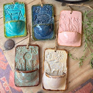 May include: Five rectangular ceramic wall planters with hanging twine. Each features a relief design of animals and flowers, including a donkey, armadillo, rabbit, windmill, and cow, in turquoise, blue, pink, sage green, and cream glaze colors.