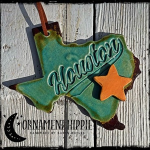 May include: A teal green ceramic Texas state ornament with the word "Houston" in white lettering. A small orange star is attached to the ornament. The ornament is hanging from a brown leather cord.