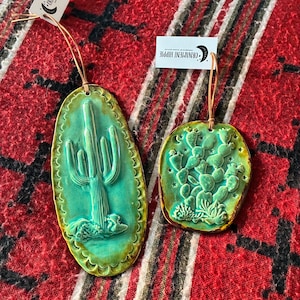 May include: Two ceramic ornaments with a southwestern theme. One is an oval shape with a cactus design, and the other is a round shape with a prickly pear cactus design. Both are turquoise with a gold trim and a brown string.