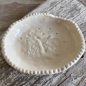 May include: A white ceramic dish with a raised design of a tree and stars. The dish has a scalloped edge.