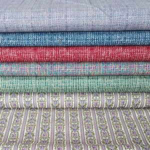 May include: A stack of folded fabric bolts featuring various solid colours including light blue, dark blue, red, teal, and green with a textured weave, topped with a piece of white fabric featuring vertical purple and green floral stripes.