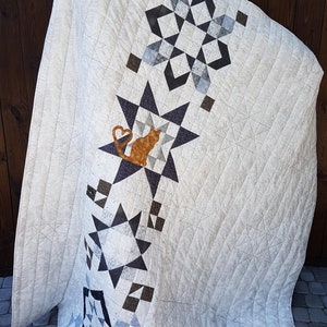 Peut inclure: Une courtepointe crème avec un motif d'étoiles géométriques dans les tons de gris, noir et marron. La courtepointe présente un appliqué de chat avec un cœur. Le texte "Example of custom quilt made to order" est visible.