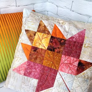 May include: Two decorative throw pillows. One pillow has a vibrant orange and green striped pattern. The other pillow features a quilted maple leaf design in shades of orange, red, and brown on a cream background.