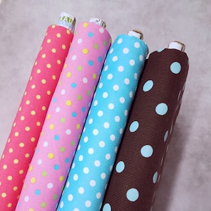 Puede incluir: Cuatro rollos de tela con diferentes patrones de lunares. Las telas son rosa con lunares amarillos, rosa con lunares verdes y amarillos, azul con lunares blancos y marrón con lunares azules.