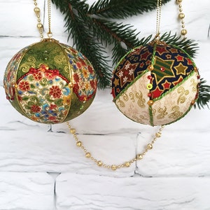 Può includere: Due decorazioni natalizie. Una è verde con motivi floreali e dettagli rossi. L'altra è un mix di tessuti blu navy, oro e crema con disegni a stella e volute. Entrambe sono impreziosite da perline e cordoncini dorati.
