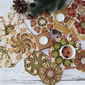May include: A selection of quilted Christmas coasters with festive designs. The coasters display patterns in red, green, and gold. A wooden candle holder with a lit candle, pine cones, and greenery are also visible, creating a seasonal display.