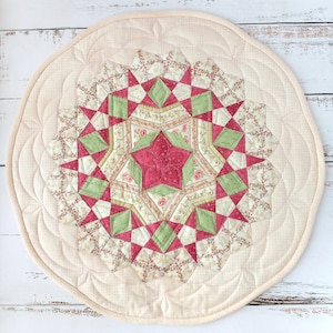 May include: A circular quilted placemat with a central starburst pattern. The starburst design incorporates red, green, and floral fabric pieces. The outer edge is a plain cream colour with decorative stitching, giving a vintage, handcrafted feel.