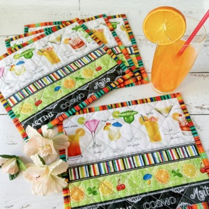 Può includere: Set di tovagliette trapuntate a tema cocktail con bordi colorati. Il tessuto bianco presenta illustrazioni di vari cocktail, tra cui martini e mojito, con dettagli di frutta e guarnizioni. È presente anche un bicchiere di succo d'arancia con cannuccia e fetta d'arancia.