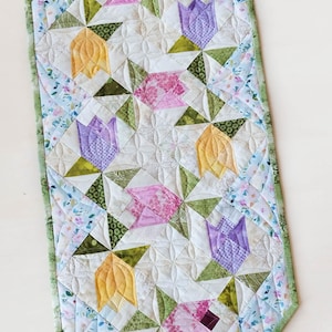 May include: A quilted table runner featuring a floral tulip design. The runner has a white center with yellow, pink, and purple tulips, green leaves, and a light blue floral border. The runner is approximately 30 inches long.
