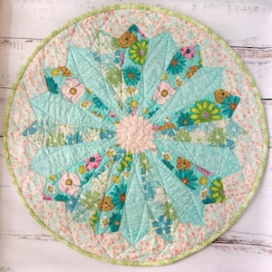 May include: A circular quilted placemat with a floral pattern. The centre has a light pink flower shape, encircled by turquoise and floral patterned segments. The outer edge has a pink and white dotted pattern, with a green border.