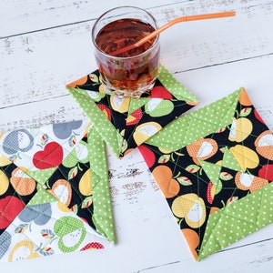 May include: Set of three quilted fabric coasters with fruit patterns. Each coaster features a black background with apples, pears, and hearts in red, yellow, and gray. The edges are trimmed with green polka dot fabric. A glass of iced tea with a straw sits on one.