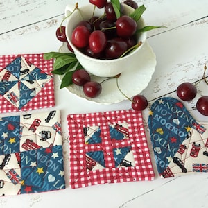 May include: Four red and white checkered fabric coasters with a London theme. The coasters feature a red double-decker bus, a Union Jack flag, and the words "I Love London". A heart-shaped coaster with the same design is also included.